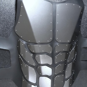 May include: A black tactical vest with a silver metallic finish and a geometric pattern of overlapping plates. The vest has a textured surface and is secured with multiple rivets.