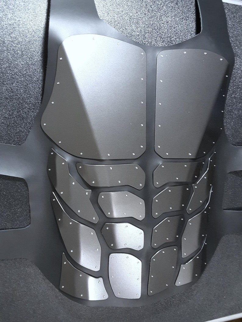 May include: A black tactical vest with a silver metallic finish and a geometric pattern of overlapping plates. The vest has a textured surface and is secured with multiple rivets.