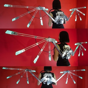 May include: A silver and black articulated wing costume with glowing green lights. The wings are attached to a backpack and can be adjusted to different positions.
