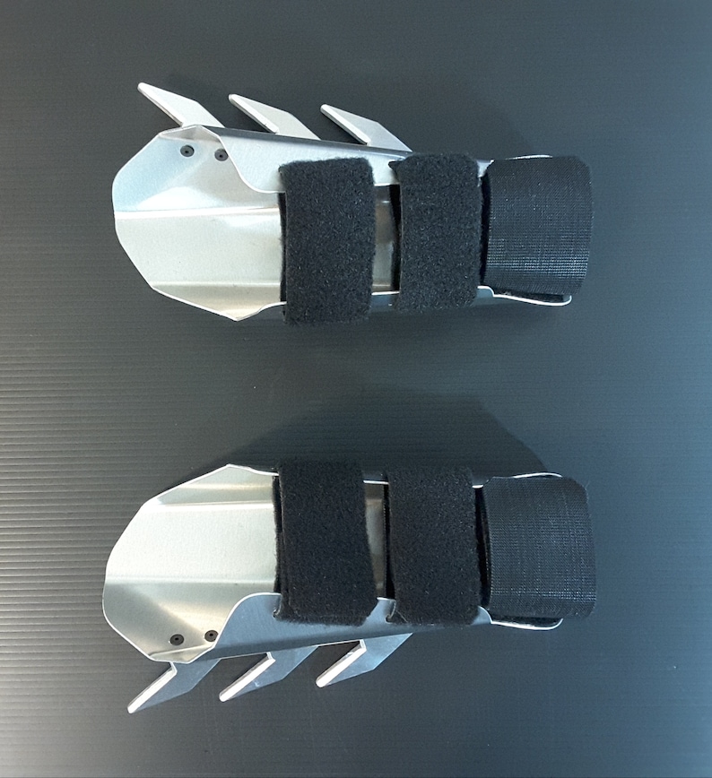 May include: Two silver metal finger guards with black adjustable straps. The guards have a jagged edge and are designed to protect fingers during activities like archery or fencing.