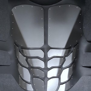 May include: A black and silver chest plate with a textured, segmented design. The chest plate is made of a flexible material and has a series of small, silver rivets.