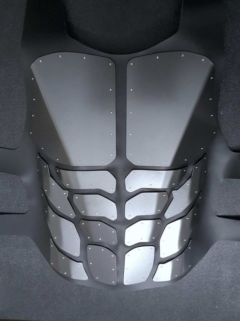 May include: A black and silver chest plate with a textured, segmented design. The chest plate is made of a flexible material and has a series of small, silver rivets.
