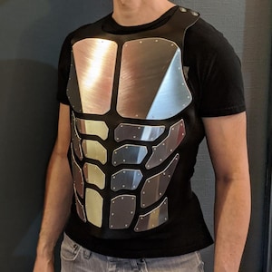May include: A black shirt with a silver, segmented, chest plate design. The plate is made of multiple pieces that resemble a human torso.