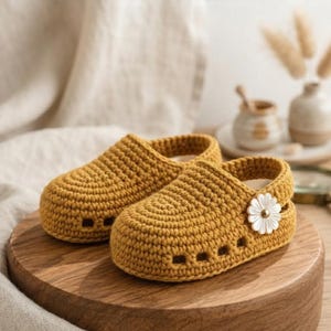 May include: A pair of mustard yellow crochet clogs featuring a rounded toe, a series of small rectangular cutouts along the sides, and a white daisy-shaped embellishment attached to the heel strap of the front shoe.