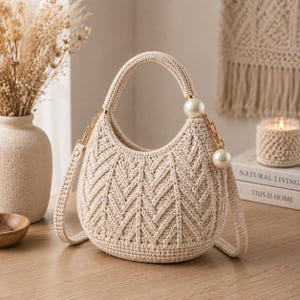 Puede incluir: Un bolso de ganchillo color crema con una forma de media luna estructurada y un patrón de espiga texturizado. El bolso incluye un asa superior, una correa de hombro desmontable y detalles de cadena dorada adornados con grandes perlas blancas.