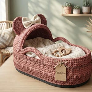 May include: A dusty rose crochet bassinet with a hood featuring two rounded ears and a cream-coloured bow. Inside lies a cream cable-knit blanket and a wooden bird-shaped rattle. A brown paper tag with the text CrochetlyStore is tied to the side.