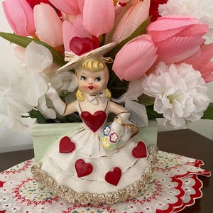 May include: A vintage figurine of a girl in a white dress with red heart accents, holding a basket of flowers, and wearing a hat. The figurine is set in a green planter with pink and white flowers. The arrangement sits on a red and white patterned doily.
