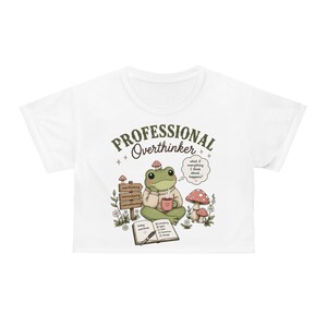 May include: A white cropped t-shirt featuring a graphic of a green frog wearing a beige jumper, holding a pink mug. The text reads "PROFESSIONAL Overthinker" above the frog, with a thought bubble saying "what if everything I think about, happens?"