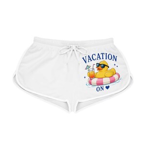 Puede incluir: Pantalones cortos deportivos blancos con cintura de cordón, con un gráfico de un pato de goma amarillo con gafas de sol, flotando en un flotador de rayas rosas y blancas mientras sostiene una bebida tropical, con el texto VACATION ON y un corazón azul.
