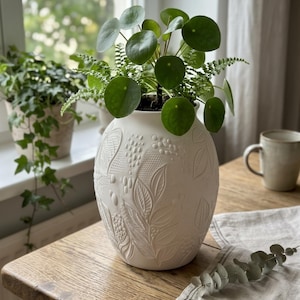 Op de afbeelding: Een witte keramische vaas met een ronde vorm en reliëfpatronen van bladeren, bessen en gestructureerde geometrische details. De vaas bevat een groene Pilea peperomioides plant met ronde bladeren en een fijne varen.