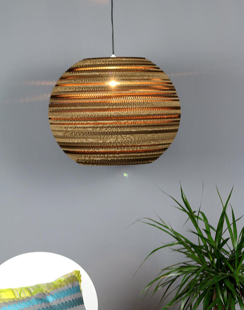 Może przedstawiać: Kulista lampa wisząca wykonana z warstwowej tektury falistej. Lampa, zawieszona na czarnym kablu, emituje ciepłe światło. Wnętrze lampy świeci. Ozdobna poduszka i zielona roślina są widoczne na pierwszym planie, na szarej ścianie.