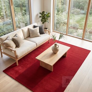 May include: A large, solid red rectangular area rug covering a light-colored wooden floor. The rug features a subtle logo in the bottom right corner with the text RUGZART. It is positioned beneath a light wood coffee table and a cream-colored sofa.