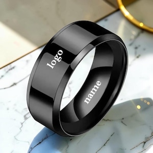 May include: A polished black ring with a smooth, reflective surface. The word logo is printed in white on the outer band, and the word name is printed in white on the inner surface of the ring, which features a wide, flat-profile band design.