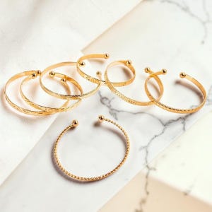 Può includere: Una collezione di bracciali a fascia in metallo color oro con design aperti e piccole estremità sferiche. I bracciali presentano varie texture, tra cui superfici lisce, motivi a corda intrecciata e dettagli finemente incisi sulle fasce.