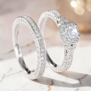 Può includere: Un set di anelli color argento composto da una fede e un anello di fidanzamento. L'anello di fidanzamento presenta una pietra centrale quadrata trasparente circondata da un alone di pietre più piccole, con altre pietre lungo il gambo.
