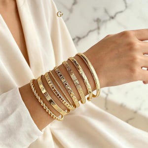 Può includere: Un set di otto bracciali rigidi in metallo color oro con varie texture e motivi, tra cui un design a corda intrecciata, motivi a stella, intagli geometrici, forme a cuore e linee incise, indossati insieme come una collezione di bracciali.