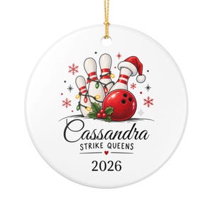 May include: A round white ceramic ornament featuring a graphic of three bowling pins wrapped in Christmas lights and a red bowling ball wearing a Santa hat. The text reads "Cassandra STRIKE QUEENS" above the year "2026" with holly accents.
