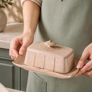 May include: A rectangular ceramic butter dish with a light beige, glazed finish. The lid features vertical embossed lines and a small, circular knob with radiating patterns. The base has a slightly flared, wavy rim with a matching beige glaze.