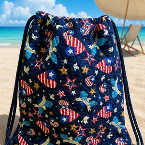 May include: A quilted drawstring backpack in navy blue featuring a repeating pattern of red, white, and blue heart-shaped flags, gold stars, and light blue eagles. The bag includes thick navy blue rope straps and gold-toned metal grommets.