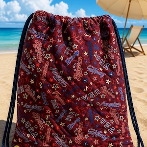 May include: A quilted drawstring backpack in deep red fabric featuring a repeating pattern of blue and white stars, along with text including USA, America, Liberty, and Freedom. The bag has thick navy blue rope straps and gold-toned metal grommets.