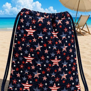 Può includere: Uno zaino trapuntato con chiusura a coulisse, caratterizzato da una base in tessuto blu scuro con un motivo ripetuto di stelle a strisce rosse e bianche e stelle bianche più piccole. La borsa include spessi cordini intrecciati blu scuro e occhielli in metallo color ottone.