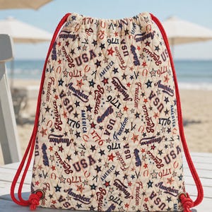 May include: A quilted drawstring backpack featuring an off-white fabric base with a repeating pattern of red and blue stars, the text USA, America, Freedom, Liberty, and 1776. The bag includes thick red rope straps threaded through metal grommets.