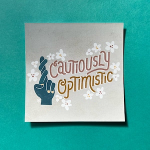 May include: A beige card with the text "Cautiously Optimistic" in brown and gold lettering. A blue hand with crossed fingers is in the center of the card. White flowers surround the text and hand.