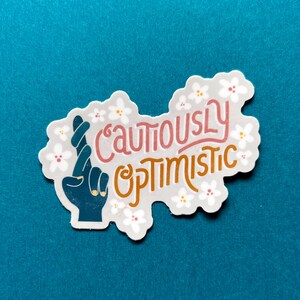 May include: A white sticker with a hand making a "fingers crossed" gesture. The sticker has a light gray background with white flowers and the text "Cautiously Optimistic" in pink and orange.