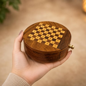 May include: A round wooden box featuring a checkerboard pattern on the lid with alternating light and dark squares, each containing a small hole. The box is crafted from dark brown wood and includes a small brass knob on the side for opening.