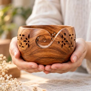 Puede incluir: Un cuenco de madera redondo para lana con un patrón de veta de madera marrón natural y un acabado liso. El cuenco tiene un distintivo corte en espiral en la parte frontal para pasar el hilo, complementado con cortes decorativos florales.