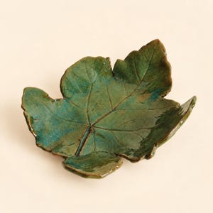 May include: A ceramic dish shaped like a maple leaf with a glossy finish. The surface features a deep green and teal glaze with visible dark brown veining that mimics the natural structure of a leaf, creating a textured and organic appearance.