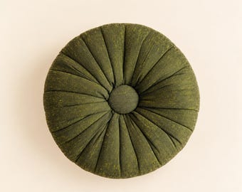 May include: A round, olive green cushion featuring a tufted design with radiating pleats and a central fabric-covered button. The surface has a textured appearance, and the overall shape is circular, creating a uniform, symmetrical look.