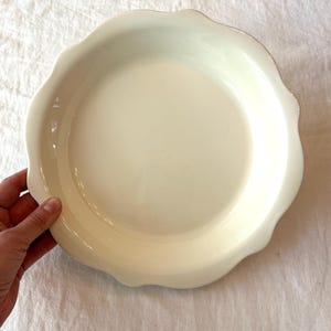 May include: A round, off-white ceramic pie dish featuring a smooth, glossy finish and a decorative scalloped edge. The dish has a wide, shallow basin and a curved rim, providing a clean, neutral appearance for baking and serving various food items.