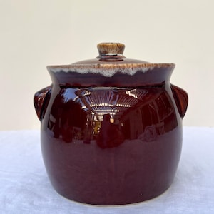 May include: A round, dark brown ceramic crock with a matching lid. The pot features two side handles and a glossy finish, with a distinctive drip-glaze effect in lighter tan and white tones along the upper rim and the top of the lid knob.