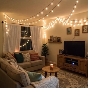 May include: A living room featuring a beige sofa with assorted throw pillows, a wooden coffee table with a candle, a wooden media console with a television, and white curtains. Strands of warm white string lights are draped across the ceiling.