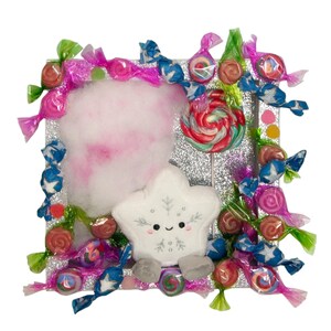 Puede incluir: Un marco cuadrado de purpurina plateada decorado con varios caramelos envueltos de colores y una piruleta grande de remolinos rojos, verdes y blancos. Una estrella de peluche blanca con un copo de nieve bordado y una cara sonriente.