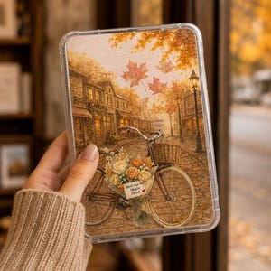 以下が含まれることがあります： 秋の石畳の通りに花かごを乗せた自転車が描かれた透明なスマホケース。紅葉したカエデの葉が舞い、街灯が並ぶ風景の中に「Meet me on Maple Street」と書かれた看板が添えられています。