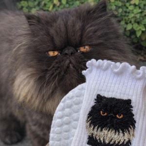 May include: A white ribbed sock featuring a ruffled top edge and an embroidered patch of a long-haired black cat with orange eyes. The cat has a dark face and a lighter, cream-colored chest area, matching the appearance of the cat behind it.