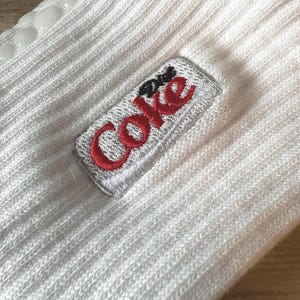 May include: White ribbed socks with a Diet Coke logo embroidered in red and silver. The socks have a non-slip grip on the sole. The socks are made of a soft, ribbed material.