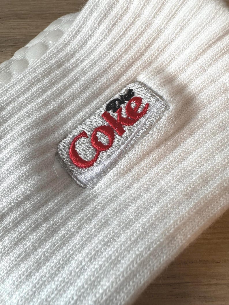 May include: White ribbed socks with a Diet Coke logo embroidered in red and silver. The socks have a non-slip grip on the sole. The socks are made of a soft, ribbed material.
