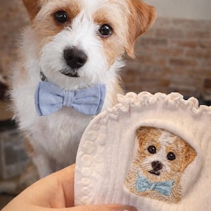 May include: A white sock featuring an embroidered portrait of a small, scruffy, tan and white dog wearing a light blue bow tie. The embroidery is framed by a ruffled edge, and the sock has a textured grip pattern on the side of the fabric.