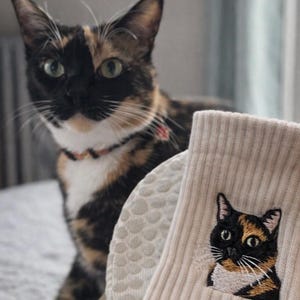 May include: A pair of beige ribbed socks featuring a detailed embroidered patch of a calico cat. The embroidery depicts a cat with black, orange, and white fur, green eyes, and white whiskers, matching the appearance of the calico cat nearby.