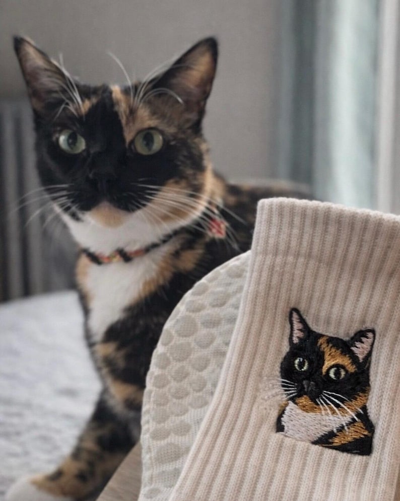 May include: A pair of beige ribbed socks featuring a detailed embroidered patch of a calico cat. The embroidery depicts a cat with black, orange, and white fur, green eyes, and white whiskers, matching the appearance of the calico cat nearby.