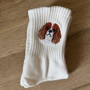 May include: A pair of white ribbed crew socks featuring a detailed embroidered patch of a Cavalier King Charles Spaniel dog face. The embroidery displays the dog's characteristic brown and white fur, long ears, and dark eyes on the upper ankle section.
