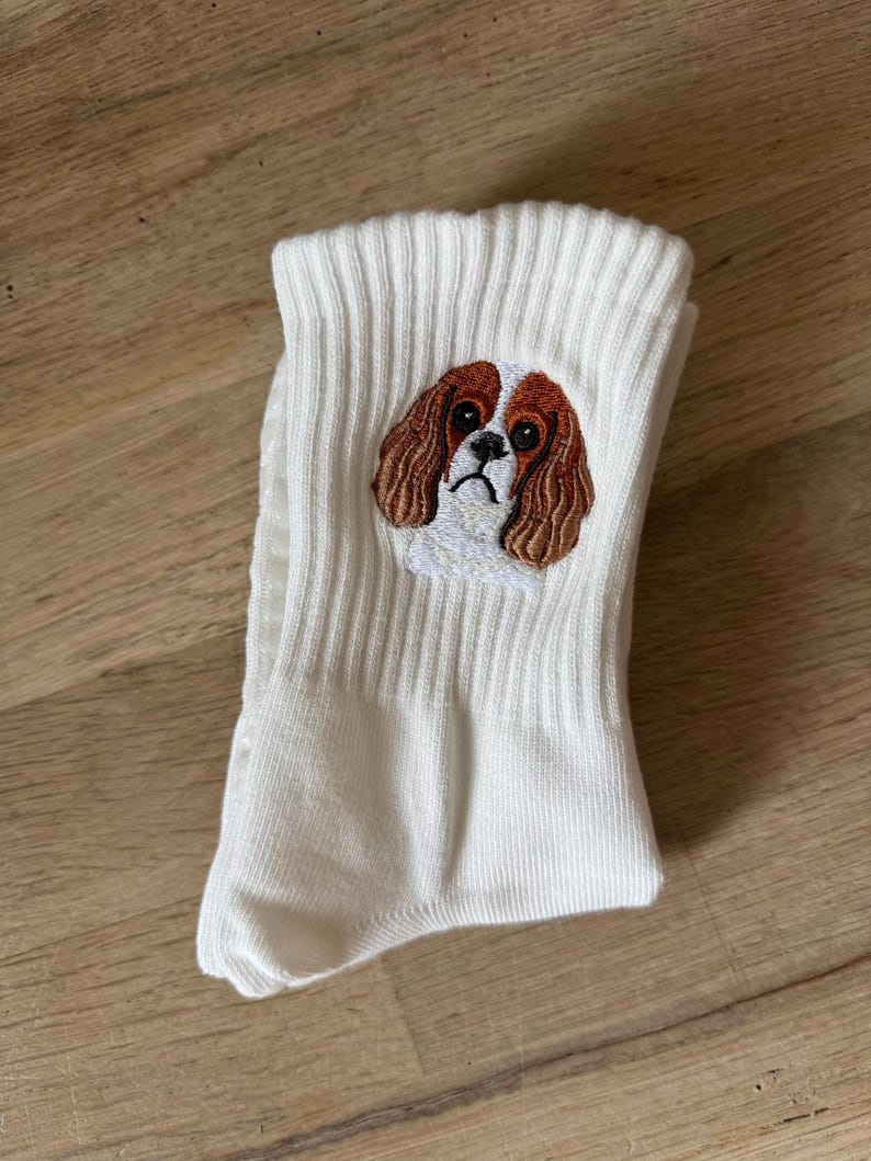 May include: A pair of white ribbed crew socks featuring a detailed embroidered patch of a Cavalier King Charles Spaniel dog face. The embroidery displays the dog's characteristic brown and white fur, long ears, and dark eyes on the upper ankle section.