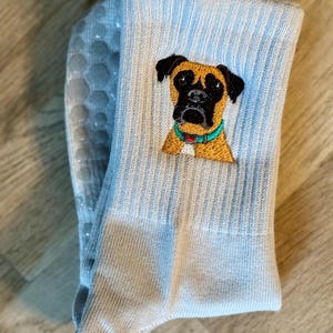 May include: A light blue crew sock featuring an embroidered portrait of a brown and black boxer dog wearing a teal collar. The sole of the sock is visible, displaying a pattern of light gray, circular, non-slip silicone grip dots.