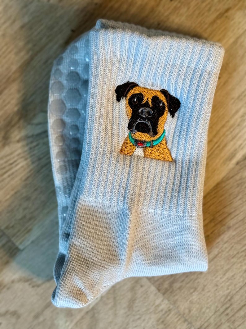 May include: A light blue crew sock featuring an embroidered portrait of a brown and black boxer dog wearing a teal collar. The sole of the sock is visible, displaying a pattern of light gray, circular, non-slip silicone grip dots.