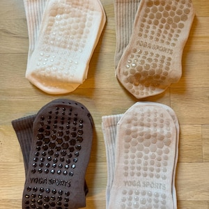 May include: Four pairs of yoga socks in neutral colors: cream, tan, grey, and light beige. Each sock has a non-slip grip pattern on the sole and the words "YOGA SPORTS" printed on the sole. The socks are made of a ribbed knit material.