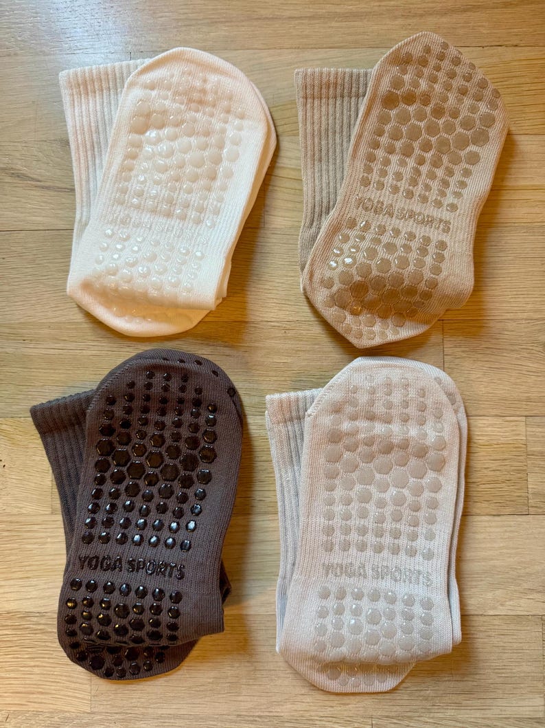 May include: Four pairs of yoga socks in neutral colors: cream, tan, grey, and light beige. Each sock has a non-slip grip pattern on the sole and the words "YOGA SPORTS" printed on the sole. The socks are made of a ribbed knit material.