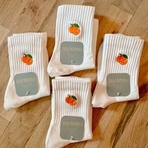 May include: Four pairs of white ribbed socks with an embroidered orange fruit design. Each sock has a tag that reads "PARKER JUNE". The socks are arranged on a wooden surface.